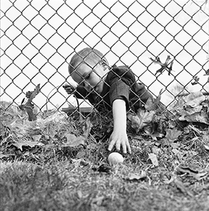 Photo of child in Sunday clothes holding Easter basket, reaching through fence to pick up egg on other side