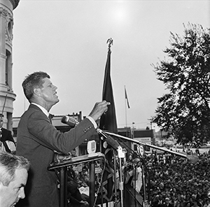 Photo of JFK at podium, surrounded by microphones, pointing while talking to crowd