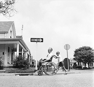 Photo of young boy pushing man in wheelchair, the opposite direction of a street sign that says "One Way"