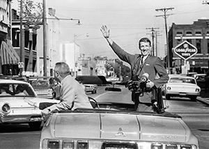 Sonny Brown riding in convertible car, standing and waving while holding a camera in his hand