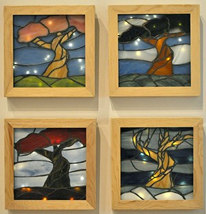 a series of 4 stained glass compositions, each with a tree and sky, ground-- all different hues. One tree has no leaves and the ground is white. Each stained glass composition is backlit with string lights- Kate Ryder, Deciduous Luminaries