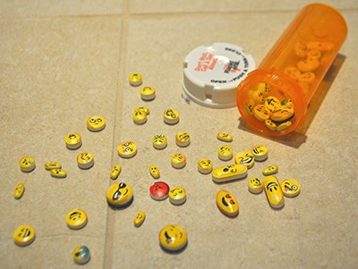 What looks like various sized pills spilled out, each with a different emoji printed on them representing different emotions. The pills have spilled out of a prescription bottle, which still has many in them. Laicee Blackwell, Daily Meds