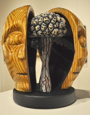 Wooden sculpture of face cut in half vertically, slid open with "brain" and brain stem visible inside. Brain lumps are made up of different happy, sad, scared, mad faces. Tyler Glendon, Tiny Monsters