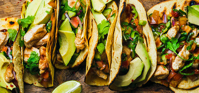 tacos all lined up with various toppings such as avocado, chicken, lime, salsa, cilantro