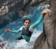 woman dangling from cliff with one hand, with intense ocean waves directly below her